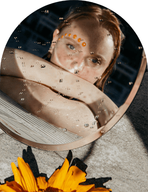 Reflection of a woman with decorative face dots in a mirror, surrounded by water droplets and flowers, emphasizing beauty and serenity.