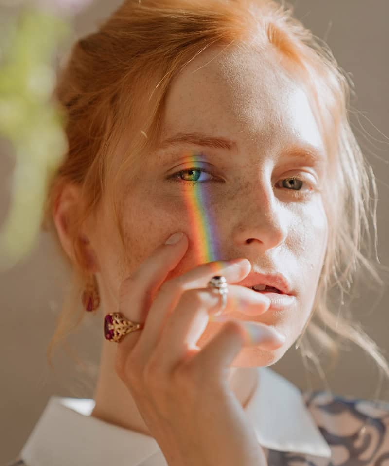 Raahni, woman with red hair and natural freckles, rainbow light reflection on face, artistic portrait photography, self-love concept, vibrant and creative image.