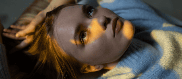Vibrant red-haired woman lying down with soft lighting on her face, cozy sweater, and contemplative expression.